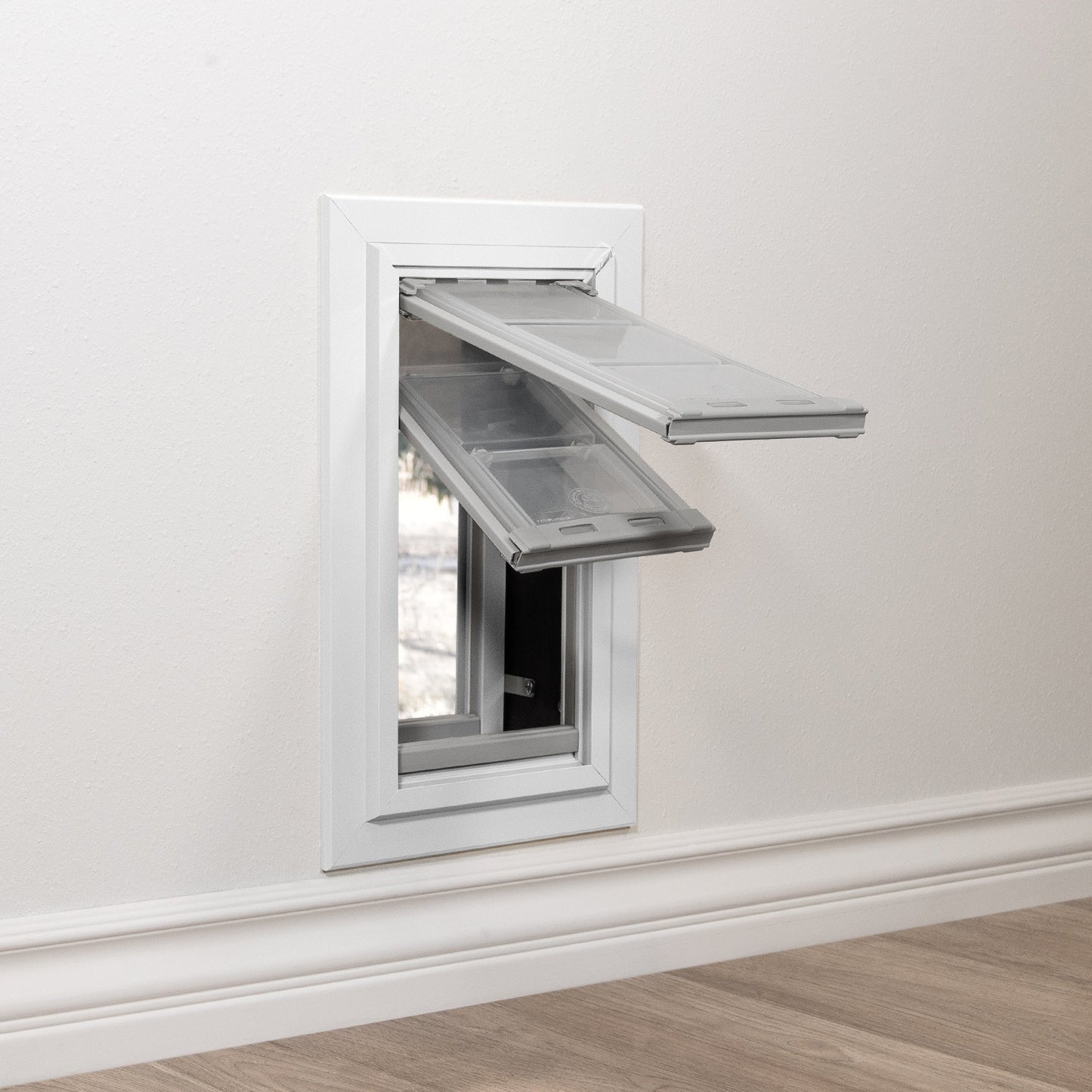 A white pet door is installed in a wall with two transparent flaps partially open.
