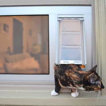 A tortoiseshell cat is partially through a pet door installed in a window.