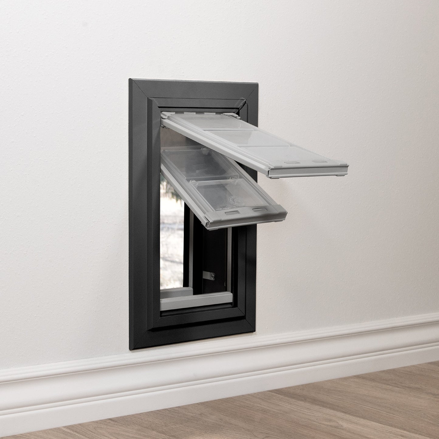 A black-framed pet door with two clear, angled flaps is installed in a white wall above a white baseboard.