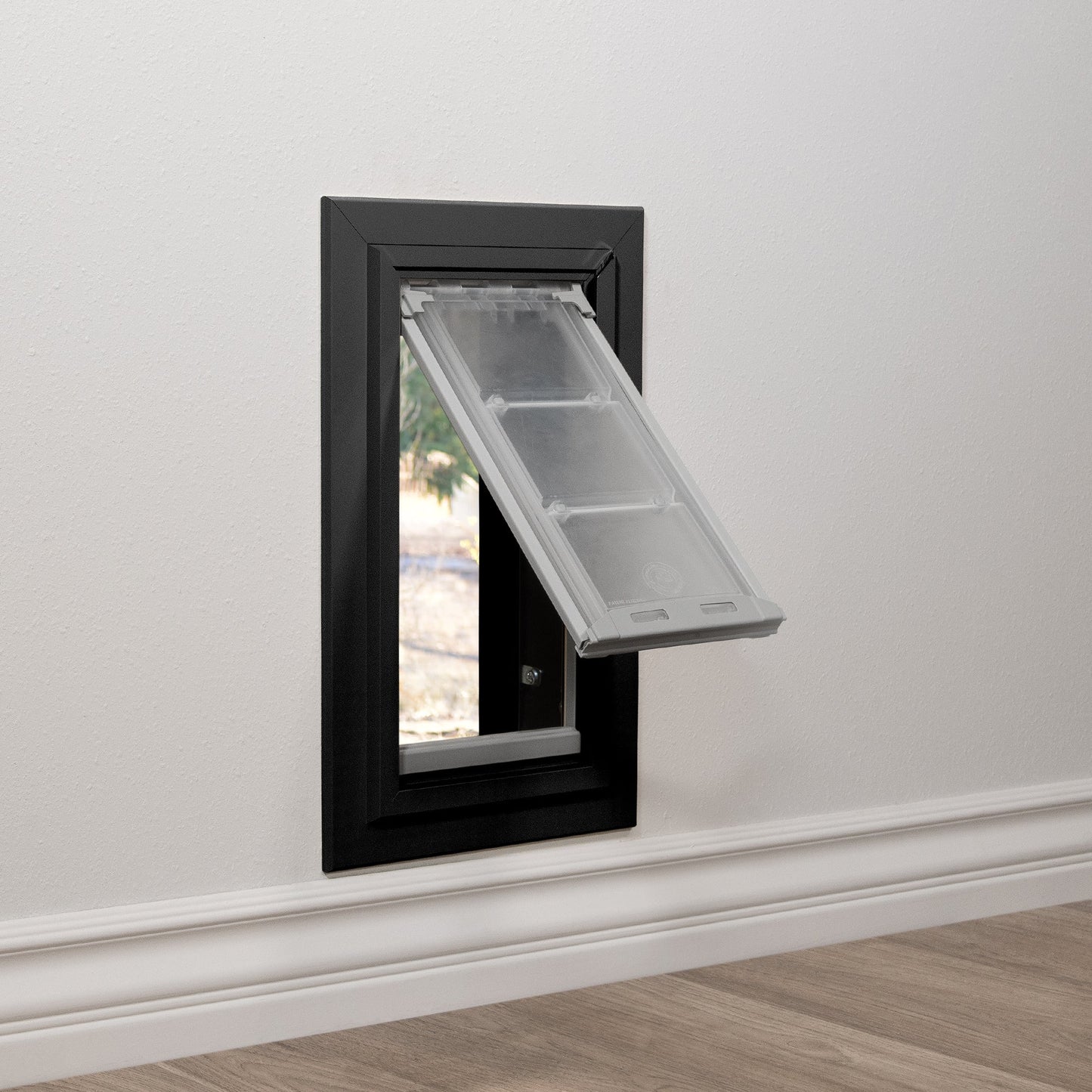 A black-framed pet door is installed in a white wall, with the clear flap partially open.