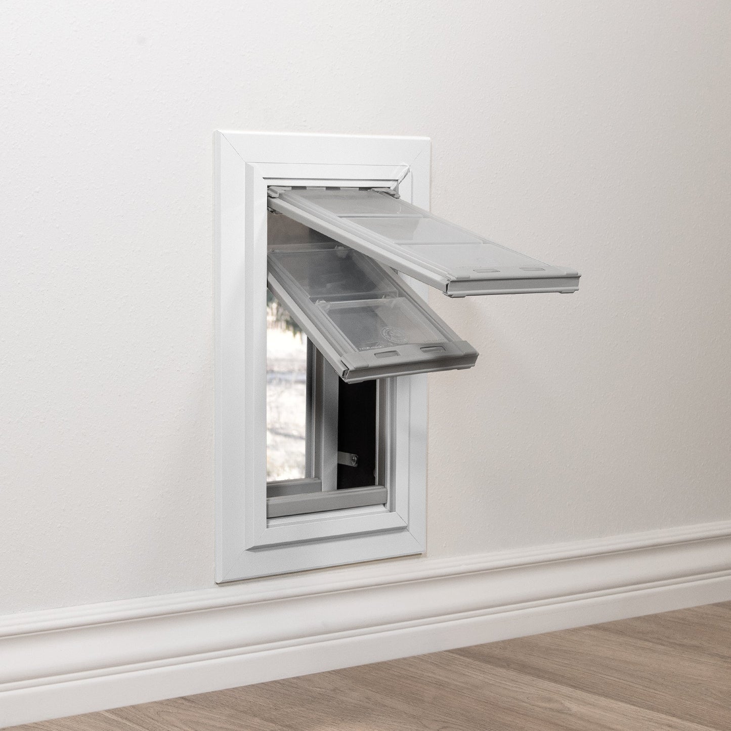 A pet door with two open flaps is installed in a white wall above a baseboard.