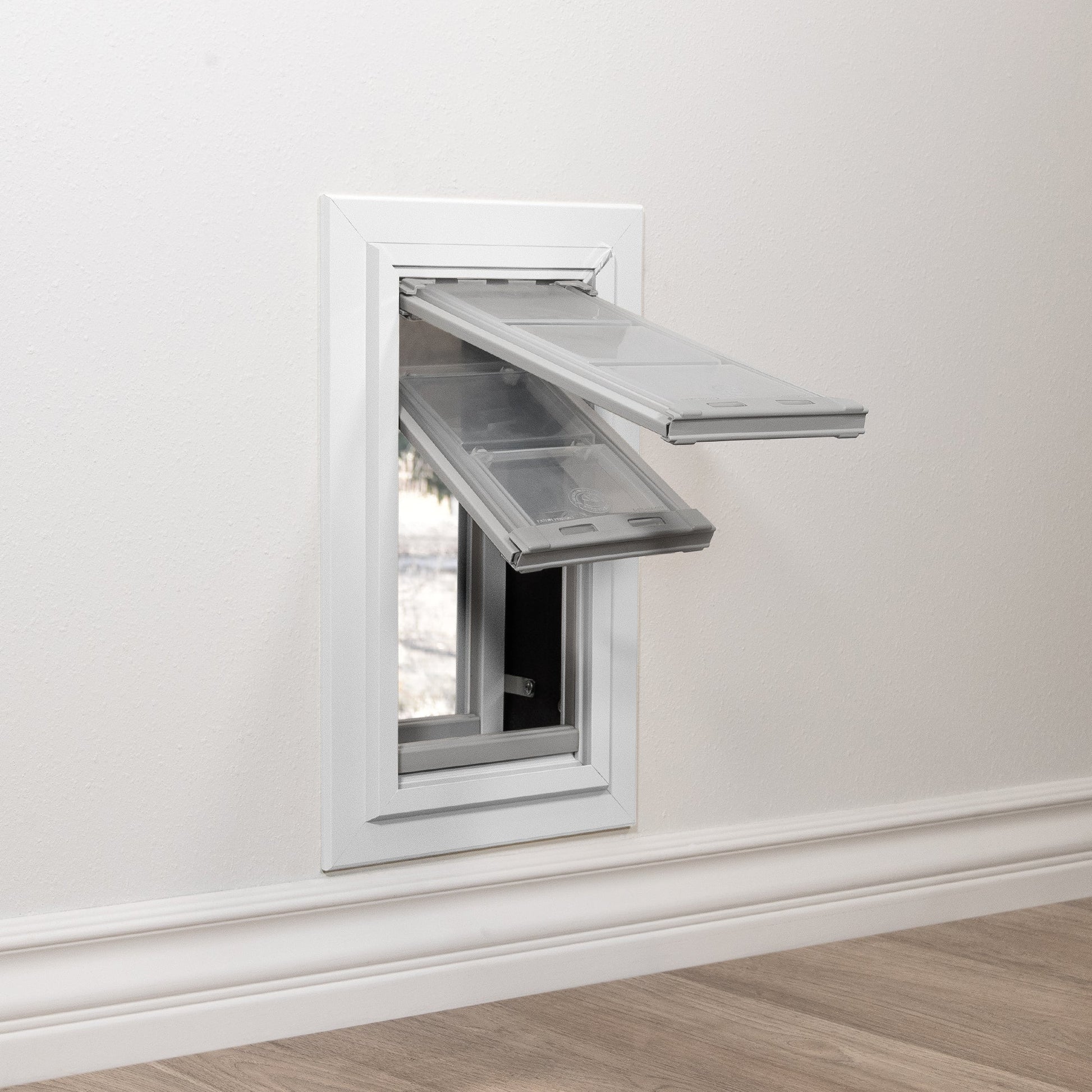 A pet door with two open flaps is installed in a white wall above a baseboard.