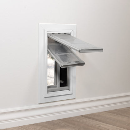 A pet door with two open flaps is installed in a white wall above a baseboard.