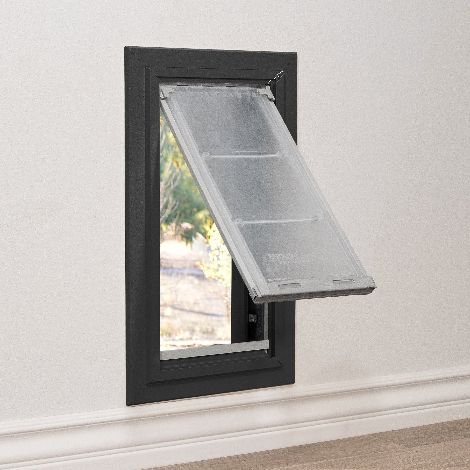 A black-framed pet door is installed in a wall, revealing an outdoor view through its partly-open, flexible flap.