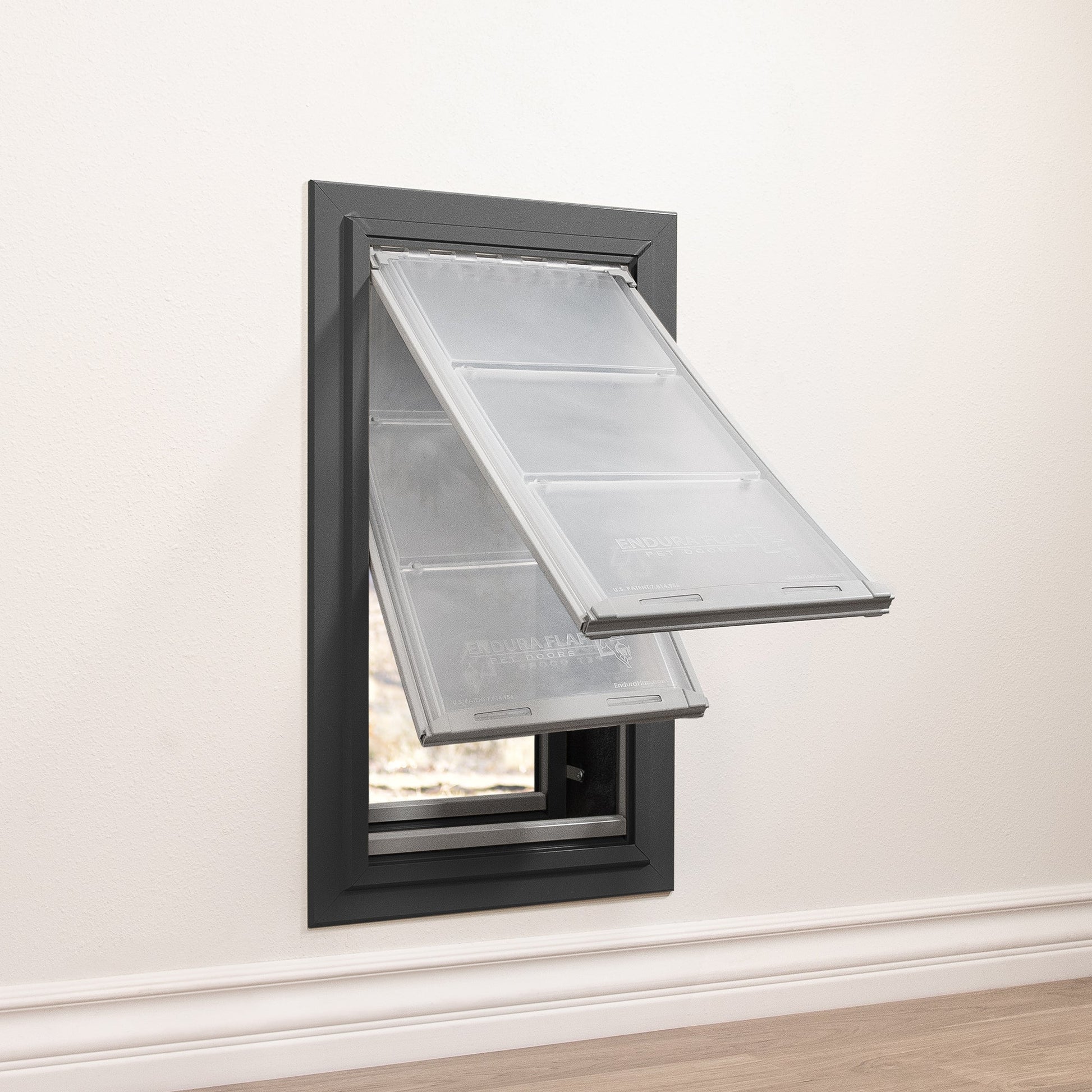 A gray-framed pet door with two partially opened clear flaps is installed in a white wall.